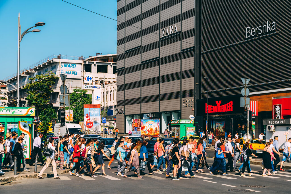 Runsas joukko kävelemässä suojatien yli. Taustalla kauppakeskus missä Zara, Bershka, New Yorker ja KFC. Yhdistelmä brutalismia, kaupallisuutta ja aurinkoista keliä.