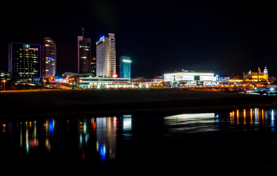 Yöaika. Casino Olympic ja muita rakennuksia. Suurelta näyttävä joki (Mahdollisesti Neris) etualalla.
