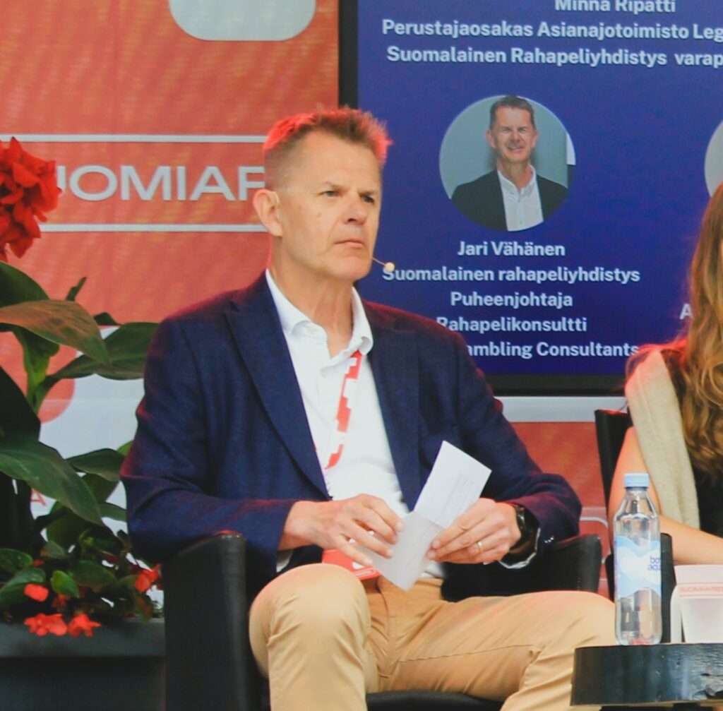 Jari Vähänen panelistossa Suomi Areena 2026 tapahtumassa. Taululta näkyy että samassa paneelissa Minna Ripatti. Vieressä toinen nainen. Vähäsen titteli on Suomalaisen rahapeliyhdistyksen puheenjohtaja ja rahapelikonsultti Finnish Gabmling Consultants.